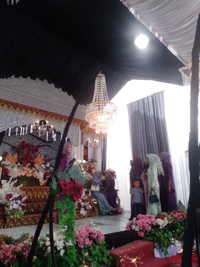 The image depicts a formal event, likely a wedding or celebratory gathering, taking place in Matangbaroh, Indonesia, beneath a large, elaborately decorated tent.

On the left side of the frame, a grand stage is set, featuring an ornate white and gold backdrop adorned with intricate carvings and a traditional gold and black patterned fabric border. Two chandeliers, one with multiple "candle" lights and another with hanging crystal teardrops, are integrated into the stage decor. Substantial floral arrangements, comprising red, orange, white, and green flowers, are displayed at the stage's base. A woman wearing a purple long-sleeved outfit and a beige hijab stands on the stage, positioned to the left, looking towards the right, behind a dark vertical stand.

A prominent, multi-tiered crystal chandelier hangs from the tent's ceiling in the upper center. The tent ceiling is draped with black fabric on the left and white fabric on the right. A bright, circular light source illuminates the upper right section of the white ceiling. Dark grey curtains serve as a backdrop on the far right of the scene.

A thick black structural pole runs vertically in the mid-ground. To the right of this pole, a group of approximately seven individuals is present. The group mainly consists of women wearing hijabs and long, formal dresses in varying shades of purple, light green, and light blue. A woman in a light green dress and matching hijab stands centrally within this group, holding a small black handbag. To her immediate left, a young boy, estimated between 5 and 7 years old, wears a grey t-shirt and dark pants, looking towards the left side of the frame. Other women in purple and light blue attire are either standing or partially seated on ornate white chairs further back.

The foreground on the right displays additional floral arrangements, predominantly featuring pink roses and white flowers, interspersed with delicate string lights. A red-covered platform is also visible in the lower right corner of the frame. The overall setting conveys a festive and ceremonial ambiance.