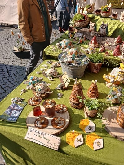 This image captures a vibrant market stall in Homburg, Germany, set on a cobblestone surface under natural daylight. The primary focus is a table draped with a textured green tablecloth, displaying numerous handcrafted items.

A person, partially visible on the left, stands facing away from the viewer, wearing a brown jacket, a dark green shirt, and dark pants, with hands in pockets. In the background, other indistinct individuals move through the market.

The stall itself is laden with a variety of decorative objects. In the foreground, on the left, a folded brochure displays images of bee-themed items. Adjacent to it is a handwritten sign on light cardstock, reading "BEE PROTECTED and Bee Happy," with "DECKEL 22,-" inscribed below. Three hexagonal yellow boxes, each featuring a "Bee Happy" logo with a bee illustration, are arranged nearby.

Towards the center, a large circular wooden slab serves as a display for four round, flat objects, likely coasters, adorned with honeycomb patterns and small bee figurines. A small glass jar containing a reddish liquid, possibly honey, and a taller cylindrical container are also present on this slab.

Further back on the table, two prominent beehive-shaped sculptures crafted from coiled material are displayed, alongside a small potted plant with ivy. Another wooden slab holds a similar beehive-shaped item.

The mid-section of the table showcases numerous decorative turtles of varying sizes, crafted from ceramic or stone, many featuring mosaic-like patterns. Some are placed within a galvanized metal bucket filled with water, while smaller turtle figurines are scattered around.

On a tiered display covered by the green cloth in the background, additional handcrafted items include several owl figurines, planters with green foliage, and small conical decorative trees. A partially visible sign in German reads "Kreativ-B...". The overall impression is that of an artisan or craft market stall.