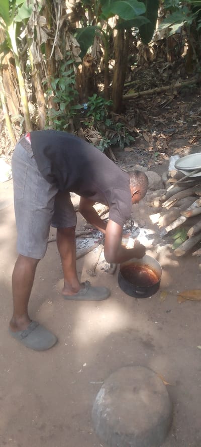 In an outdoor, unpaved setting in Ohafia, Nigeria, a male individual is depicted from a slightly elevated perspective, bent forward at the waist. The subject, wearing a black short-sleeved t-shirt, grey shorts, and light grey slip-on slippers, is actively tending to a cooking pot. A red and black patterned underwear waistband is visible above the shorts. His arms are positioned over a dark metal pot containing a reddish-brown substance, which is cooking over an open wood fire. The fire consists of glowing embers and ash, surrounded by various logs and wood debris. To the right of the cooking area, a stack of unburnt logs is visible, along with a silver-colored metal tray or lid. The ground is bare earth, light brown in color, with scattered organic matter. In the foreground, a large, rounded grey stone or concrete block rests on the ground. The background features dense, natural vegetation, primarily consisting of banana plants with large green leaves and dry, brown fronds, indicating a lush, semi-rural environment under natural light.