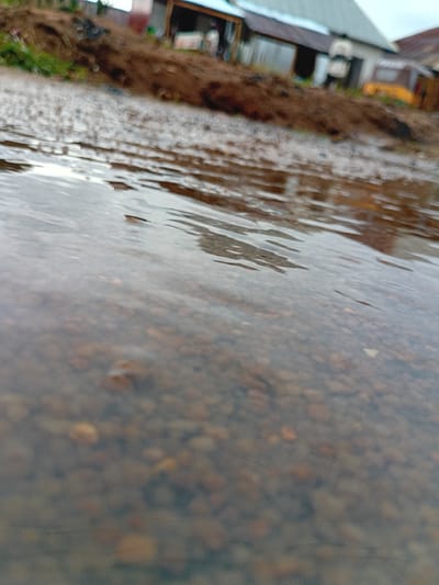 This media file displays a close-up, angled view of a water puddle on a surface that appears to be a road or pathway, with small stones visible beneath the water. The water is rippled, suggesting it might be raining or there is movement of water. To the right of the puddle, there is a rough, textured surface that transitions into a muddy embankment with patches of green vegetation. Further to the right, structures resembling buildings or stalls are partially visible, with some metal sheeting and wooden elements. A yellow object, possibly a vehicle or part of construction equipment, is also discernible in the background. The scene appears to be outdoors, with the lighting suggesting it could be daytime, possibly overcast or after rain. There are no people or explicit human activity directly captured in the frame, though the presence of buildings suggests a populated area. The overall impression is of a damp, possibly rural or semi-urban environment in Nigeria. No text is visible.