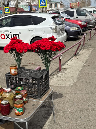 The image depicts a street scene in Borzya, Russia. In the foreground, a metal cart is laden with two black plastic crates, numerous glass jars filled with preserved food, and two large bunches of red carnations. The cart appears to be stationary on the side of the road.
In the background, several vehicles are parked along the street, including a white van with a cardboard box on its roof, a red car, and a white car displaying the word "Maxim" in large red letters and Russian text. A pedestrian crossing sign is visible near the vehicles. There are also some red metal barricades along the street. The surrounding environment features buildings, trees, and a paved road. The overall scene suggests an impromptu roadside market or sale.