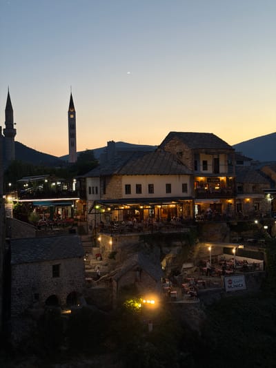 This is a twilight photograph of the Old Town of Mostar, Bosnia and Herzegovina, taken from an elevated vantage point looking down onto a series of restaurants and historic stone buildings. Two prominent minarets are visible in the background, one to the left and another taller, more slender one to the right, both silhouetted against the pale orange and blue twilight sky.
The foreground and midground are dominated by multi-level stone structures with pitched, dark-tiled roofs. These buildings appear to be part of a hillside cascading down towards a river, though the river itself is not visible. The structures are illuminated by a warm, artificial light emanating from within and from lamps strategically placed on terraces and walkways.
Several outdoor dining areas are set up on these terraces, populated by numerous patrons seated at tables. The tables are covered with white or red tablecloths, and some are adorned with flowers. Waitstaff are visible serving customers. One sign reads "konoba MLINICA" in large white letters on a red background.
The architecture is rustic and traditional, with visible stone masonry and wooden accents. Steep, stone staircases wind between the levels, also lit by lamps. The hillside is partially covered in dense green foliage, which appears as dark masses in the fading light. The overall impression is one of a lively, historic urban setting bathed in the soft glow of evening light.