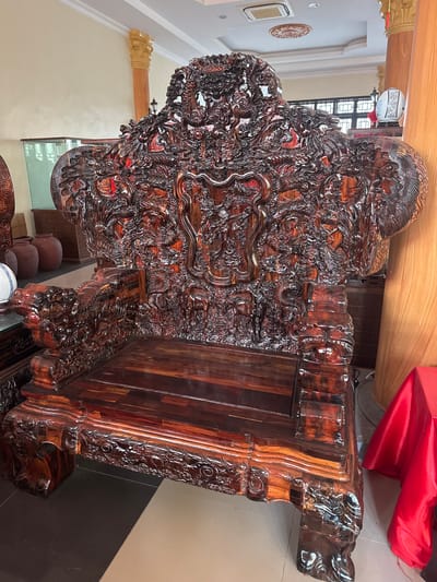 This is a medium-shot, eye-level, full-length photograph of an elaborately carved wooden armchair, possibly a throne, positioned indoors. The armchair is a deep, rich brown with a high, intricately detailed back featuring numerous carved dragons and other mythical creatures, all rendered in a dark, glossy wood. The seat and armrests are also heavily carved. The chair appears to be made of dark, polished wood with visible grain patterns.
The armchair is the central and dominant subject of the image. To the left of the armchair, a portion of another wooden object with similar carving is visible. In the background, several large, reddish-brown ceramic jars are stacked. Further back, a wooden cabinet and a display case with glass doors can be seen.
The environment appears to be an indoor space, possibly a showroom or a shop. The floor is tiled in alternating dark grey and beige squares. There are also large, round wooden pillars supporting the ceiling, which is white with recessed lighting fixtures and a decorative circular element. To the far right, a bright red cloth drapes over an unseen object. The lighting is bright and even, illuminating the details of the armchair. There are no people or animals present in the image. The image is of good quality and focus, with the details of the carvings being clearly visible.
The location is stated to be in the city of Hy Cương, Vietnam.
