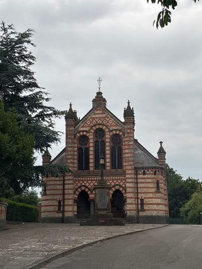 The image depicts a historical building, likely a chapel or small church, with distinctive red and white striped brickwork. The building has arched entrances, arched windows with tracery, and decorative spires topped with crosses. In front of the building, there is a stone monument or cenotaph with inscribed text. A lamppost stands beside the monument. The ground in front of the building is paved with stone slabs, and a paved road leads towards the structure. Lush green trees and hedges surround the building, suggesting a park-like or cemetery setting. The sky is overcast. The building is located in the town of Winsford, United Kingdom.