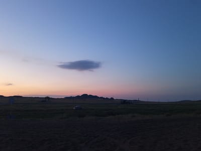 The image depicts a dusky, rural landscape, likely in the Bayanaul rural district of Kazakhstan. The sky transitions from a pale orange and pink near the horizon to a deep blue overhead, with a single, thin cloud illuminated by the setting sun. The terrain is predominantly flat, covered in dark green grass. In the mid-ground, a car is visible on the left, and further to the right, what appears to be a truck or a large vehicle is present. Several utility poles are scattered across the field, marking the presence of infrastructure. The foreground is a dark, rough, and textured surface, possibly soil or sand, showing tire tracks. Hills or low mountains are visible in the distance on the horizon, silhouetted against the fading light.