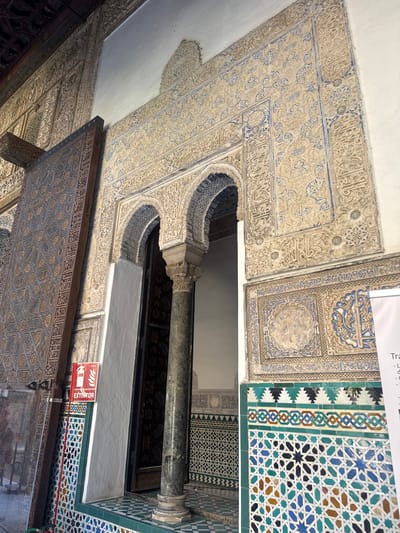 The image shows an interior view of a building with intricate architectural details, likely a historical site in Seville, Spain. The walls and ceiling are adorned with elaborate stucco work featuring geometric patterns and Arabic calligraphy in beige and blue tones. To the right, a section of wall is covered in colorful mosaic tiles in a geometric star pattern, with shades of blue, green, white, and brown.
A large, dark marble column stands in the center, supporting an archway with more detailed stucco and tile work. Below the archway, a wooden structure with carved panels, also featuring geometric patterns and calligraphy, is visible. A red and white sign with the word "EXTINTOR" (Spanish for fire extinguisher) and an icon of a fire extinguisher is mounted on the wall to the right of the column. A white paper with some text is partially visible at the top right of the image.
The lighting suggests an indoor setting, possibly well-lit by artificial lights or natural light filtering from an unseen source. The overall impression is one of rich historical and artistic craftsmanship. No people are visible in the image.