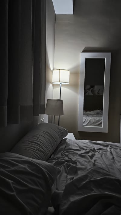 The image depicts a dimly lit bedroom in what is presumably a hotel room in Alhama de Aragón, Spain. A large, rumpled bed with white linens dominates the foreground, with a long pillow positioned at the head. To the left, dark grey curtains are drawn, obscuring a window. In the background, a tall, white-framed mirror hangs on the wall, reflecting a portion of the room. Two lamps are visible: one is a floor lamp with a white cylindrical shade, and another, smaller lamp with a conical shade, sits on a surface to the right of the bed. The lighting is subdued, casting shadows and creating a moody atmosphere. There are no people visible in the scene. The overall impression is one of a quiet, possibly untidy, private space.