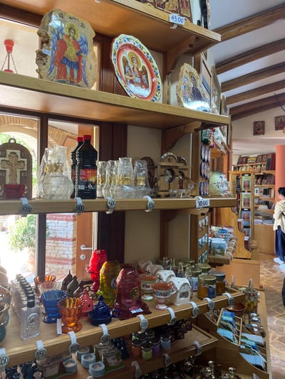 The image shows a shop with various items for sale, including glass lamps, religious icons, and bottles of oil, located in a wooden building. The shop is located in Moni Archaggelou, Greece, at coordinates 40.597 latitude and 24.702 longitude. The walls are covered in wooden shelves with various items for sale. The shelves are labeled with prices, ranging from 200 to 3,000. There are also some items for sale on a table next to the shelves. The shop is brightly lit and the shelves are well-stocked. A person can be seen in the background, looking at the items for sale. The shop is located near a window that overlooks a courtyard with stone walls and trees. The items for sale include small glass lamps, ornate religious icons, bottles of oil, and other religious paraphernalia. The scene is peaceful and serene, suggesting a place where people can purchase religious items.
