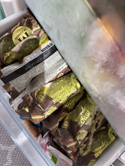A freezer drawer, situated in the city of Haßloch, Germany, is densely packed with multiple individually wrapped Magnum Signature Pistache ice cream bars. The packaging is primarily green and brown, featuring images of pistachios and chocolate, with the prominent "MAGNUM" logo and "LA PISTACHE Signature" text. Visible details on the packaging include "DIPPED IN MAGNUM CHOCOLATE," a best-before date of "11/2027" with a time stamp of "10:55," and a volume of "90 ml e / 69 g." Nutritional facts and ingredients such as "Pistazien," "Milch," "Zucker," "Kakao," "Haselnuss," "Mandel," and "CASHEW" are partially legible across various wrappers. The interior of the freezer exhibits significant frost on the right-hand wall and a white plastic divider. A small, orange, irregularly shaped food item is visible beneath some of the ice cream bars. A grey, patterned surface is discernible in the bottom-left corner of the image, outside the freezer compartment.