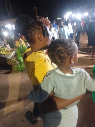 A rotated, close-up shot shows a person in a yellow shirt, holding a child in a gray sweatshirt. The child's head is near the person's face. To the left, there's a crowd of people illuminated by bright lights, and a display of green and yellow fruits, likely bananas and other produce. The ground appears to be a dirt or gravel path. The overall lighting suggests it is taken at night.