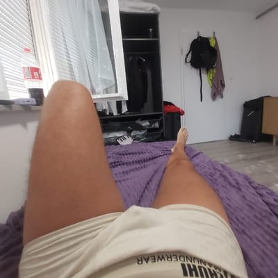 The image depicts a first-person perspective of a person lying on a bed with a purple textured blanket. The person's legs, clad in light-colored underwear with visible text "MAHNUDERWEAR," are prominently featured. The underwear has a ribbed texture. To the left, a windowsill is visible with blinds and a partially obscured bottle of soda. Further left, a white curtain hangs. In the background, a dark grey wardrobe with shelves containing various items, including clothing, is present. To the right, a white wall displays a backpack and towels hanging on a hook. A dark piece of luggage and a wooden cabinet are also visible. The floor appears to be light-colored wood. The scene is indoors, likely a bedroom. There is no specific indication that the location is in the city of Gärtringen, Germany.