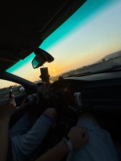The image captures the interior perspective from a vehicle traveling on a multi-lane road in Devnya, Bulgaria, during either sunrise or sunset. The sun, a distinct orange orb, is low on the horizon through the front windshield, casting intense light into the vehicle's cabin. The upper section of the windshield exhibits a green-blue tint, and the exterior surface displays numerous small specks of dust or dirt.
Two occupants are visible within the vehicle. The individual in the driver's seat, on the left, has their left hand holding a black smartphone and their right hand partially gripping a black multi-function steering wheel with a visible emblem. This person is wearing blue denim shorts or jeans. A second individual, presumed to be in the front passenger seat, has their left arm visible on the right side of the frame, wearing a white smartwatch. Their lower torso, clad in blue denim shorts or jeans, is also visible.
The vehicle's interior features a dark dashboard. A silver cylindrical object, resembling a beverage can, is situated in a console or holder on the passenger side. A rearview mirror is centrally positioned, from which a black air freshener with a white 'X' design hangs, accompanied by a miniature cartoon car and several other small decorative charms. The interior roof liner and sun visors are dark in color.
The exterior scene depicts a highway or major road extending into the distance, bordered by guardrails and sparse vegetation. Distant hills or low mountains are visible on the right horizon, and other vehicles appear as distant forms on the roadway.
