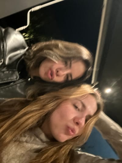 Two young women pose for a selfie, both with pursed lips. The woman on the left has short, wavy brown hair and is wearing a black leather jacket. The woman on the right has long, blonde hair and is wearing a tan knit sweater. The background appears to be outdoors at night, with a dark sky and faint light in the distance. The image has a slight blur, suggesting it was taken quickly.