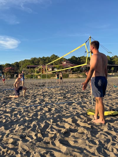 This is a medium-shot, eye-level, outdoor photograph taken on a sandy beach in Pistula, Montenegro. The scene depicts a group of people playing beach volleyball on a sunny day. The sand is light brown and textured with footprints and shadows. A bright yellow volleyball net is set up across the scene, with yellow poles supporting it. In the foreground, to the right, a shirtless man wearing navy blue shorts stands with his back to the camera, facing the net. His right arm is slightly extended, and his left hand is held up, possibly in a gesture of communication or instruction. To the left, in the mid-ground, a shirtless man in navy blue shorts and sunglasses is kneeling on the sand, looking towards the net. He has the number '3' visible on his shorts. In the background, other people are visible playing volleyball. Beyond the volleyball court, there are trees and some buildings with red roofs, suggesting a coastal resort area. The sky is a clear, bright blue with a few wispy white clouds. The overall impression is one of a casual, recreational activity on a warm, sunny day.
