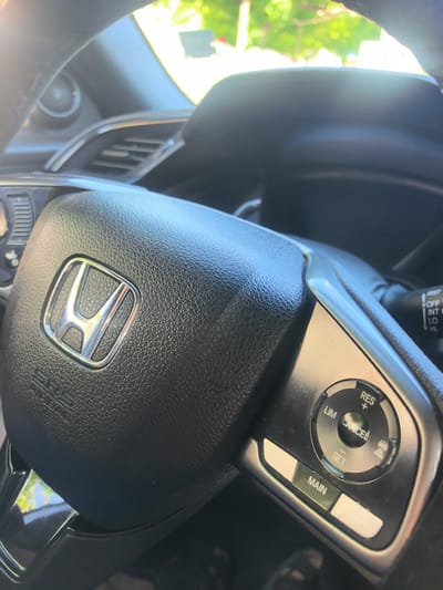 The image shows a close-up view of a car's steering wheel, specifically the driver's side. The Honda logo is prominently displayed in the center of the steering wheel. To the right of the logo are various control buttons, including "RES+", "CANCEL", "LIM", and "SET", suggesting a cruise control system. There are also "MAIN" and two blank buttons. The steering wheel appears to be made of black leather. In the background, out of focus, is a bright, sunlit environment with greenery, hinting at an outdoor setting, possibly a street or a parking lot. The sun is casting strong highlights and creating a lens flare effect in the upper left portion of the image. There are no people visible in the frame. The image is taken from the perspective of someone sitting in the driver's seat. The location in the city of Veliki Preslav, Bulgaria is not determinable from the image itself.