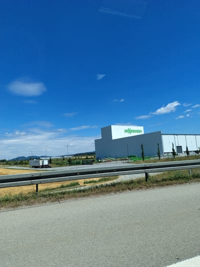 The image shows a large industrial building with the name "SENNEBOGEN" in green lettering on its facade. The building is white and has a modern design with large windows and loading docks. In front of the building, there are several pieces of green industrial equipment, possibly for construction or material handling. To the left of the building, a white semi-trailer truck is parked. The area around the building appears to be an industrial park, with flat terrain and some trees. The sky is blue with scattered clouds, indicating fair weather. The foreground of the image shows a road and a metal guardrail, suggesting the photo was taken from a vehicle driving by. This industrial facility is located in Steinach, Germany.