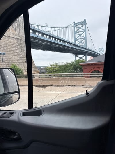 The image is a perspective view from inside a vehicle, looking out through the passenger side window towards the Benjamin Franklin Bridge in Philadelphia, Pennsylvania. The bridge, a prominent suspension bridge, dominates the upper portion of the frame, its steel structure rendered in a light blue hue against an overcast sky. The road deck of the bridge is visible, supported by intricate steel trusses. To the left of the bridge, a large, rough-hewn stone structure, likely part of the bridge's approach or support system, is visible.

In the mid-ground, a concrete barrier with graffiti marks separates lanes of traffic. Beyond the barrier, a red brick building with arched windows and a dark roof is situated to the right of the bridge's structure. Green trees and other vegetation are interspersed between the barrier and the bridge.

The foreground of the image shows the interior of the vehicle, including the dark gray plastic molding of the door panel, a side-view mirror with a reflection of traffic, and a black pillar obscuring part of the view. The overall impression is of a travel scene, capturing a well-known urban landmark from a moving vehicle.