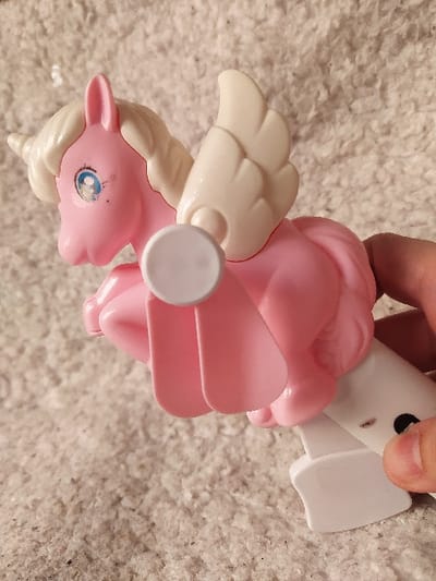 The image displays a plastic toy in Harasta, Syria. The toy depicts a pegasus-unicorn hybrid, predominantly pink in color, with a white spiraled horn, white sculpted mane, and white articulated wings featuring a feathered texture. Its eyes are blue with black pupils and a small white highlight, showing minor scuff marks near the left eye. A mechanical element is integrated into the toy's underside, consisting of a white circular component from which two paddle-shaped pink blades extend downwards. A human hand, showing a thumb and index finger, grips a white plastic base that supports the toy from below. This white base exhibits a small black circular detail and a brown speck on its surface. The background is a uniformly light-colored, textured fabric, resembling carpet or upholstery.