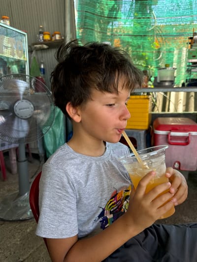 An outdoor scene, possibly in Hồng Sơn, Vietnam, captures a young boy with dark, tousled hair, wearing a grey t-shirt with a colorful graphic print on the left side of the chest. He is seated and holding a clear plastic cup filled with a yellow beverage and ice, drinking through a yellow straw. His hands are positioned around the cup, with his right hand holding it from the side and his left hand supporting the bottom.
In the background, a silver oscillating fan stands to the left of the boy, with its protective grille visible. Behind the fan, a glass display case with items inside is partially obscured. Further back, shelves hold various containers, including yellow and red plastic storage bins. A green mesh material hangs in the background, suggesting an outdoor or semi-covered area. The ground appears to be concrete. The overall impression is a casual, possibly roadside, setting.