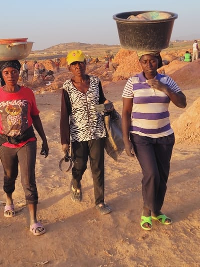 This image captures three adult women walking across a dusty, reddish-brown earthen landscape within the city of Shen, Nigeria. The background reveals numerous other individuals engaged in manual labor amidst piles of excavated earth, suggesting an outdoor work site. The sky is clear.
From left to right:
1. A woman wearing a red t-shirt with a graphic, dark cropped pants, and light pink slide sandals, balances a light-colored, empty basin on her head. She holds a dark object in her right hand.
2. A woman wearing a yellow baseball cap, a black and white zebra-print long-sleeved shirt, dark pants, and dark closed-toe sandals, carries a dark bag on her left arm and holds a dark circular object in her right hand. She looks directly at the camera.
3. A woman wearing a white and light purple/blue striped polo shirt, dark trousers, and bright green slide sandals, balances a dark bucket filled with light-colored material on her head. Her right hand is raised towards her chest, displaying light-colored dust on her fingers.
All three women have dark complexions and neutral expressions. Their primary action is walking while transporting materials or tools, indicative of physical labor in the depicted environment.