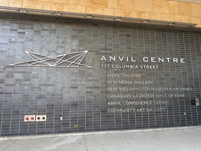 The image shows the exterior facade of the Anvil Centre located at 777 Columbia Street in New Westminster, Canada. The facade is constructed of dark grey, rectangular tiles with white lettering indicating the building's name and address. Below "ANVIL CENTRE," the text "777 COLUMBIA STREET" is displayed. Further down, a list of establishments within the Anvil Centre is visible: "ANVIL THEATRE," "NEW MEDIA GALLERY," "NEW WESTMINSTER MUSEUM & ARCHIVES," "CANADIAN LACROSSE HALL OF FAME," "ANVIL CONFERENCE CENTRE," and "COMMUNITY ART GALLERY." To the left of the text, a white abstract geometric logo is mounted on the tiled wall. In the lower left corner, two red fire department connections are visible, labeled "AUTO SPRINKLER STANDPIPE" and "MAX 150 PSI" on one, and "AUTO SPRINKLER STANDPIPE LEVEL 5 TO ROOF" and "MAX 105 PSI" on the other. A sidewalk runs along the base of the building, with a small metal plaque visible on the tiled wall near the sidewalk in the lower right quadrant. The top portion of the image reveals a beige, textured wall and a metal grate with overhead lighting fixtures. The scene is brightly lit, suggesting daylight conditions.