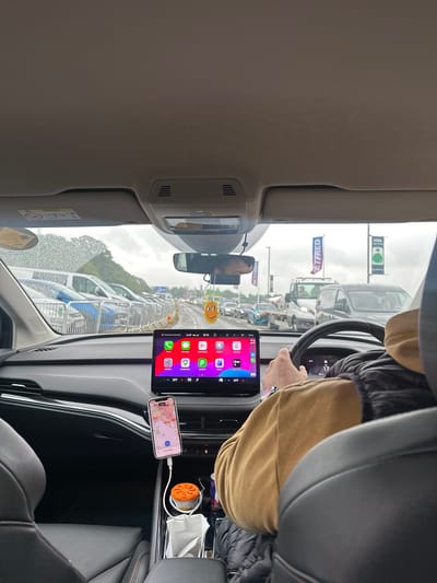 The image depicts the interior of a car, specifically the view from the front passenger seat looking forward through the windshield. A driver, identifiable by a right arm and shoulder clad in a light brown long-sleeved top and a dark puffy vest, grips the steering wheel. A watch is visible on the driver's left wrist.

The car's interior features a grey headliner, a sun visor with an "AIRBAG" warning label on the left, and a rearview mirror from which a yellow sun-shaped air freshener hangs. A large central infotainment screen displays a grid of applications including Phone, Messages, Now Playing, SKODA, Settings, Google Maps, JustPark, Teams, Uber, and Uber Driver. The top bar of the screen shows "Tattenham Corner Road" as a location and the time "18:16". A vertically mounted smartphone (iPhone) is visible on the dashboard below the infotainment screen, displaying a map interface, an "Uber Driver" app logo, and the text "You're offline". This phone is connected by a white charging cable to a port near the center console. The console area contains a white tissue box or stack of tissues, an orange cylindrical object (possibly an air freshener or diffuser) in a cupholder, and a red beverage can.

Through the rain-speckled windshield, an overcast sky and heavy traffic are visible. On the left side of the road, multiple cars, including blue and white vans, are parked behind metal crowd barriers. Trees are visible in the background on the left. On the right side of the road, several large banners are displayed, one clearly showing "JETFRED" (likely Betfred) and another with "1986". The road ahead shows more vehicles in what appears to be a queue. The location is Tattenham Corner Road, situated in Epsom and Ewell, United Kingdom, consistent with the identified road name on the infotainment system and the presence of event-related banners.