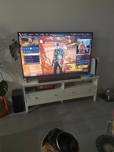 The image shows a living room in Vilanova i la Geltrú, Spain, with a large television displaying a video game interface. The game appears to be a character-based action or role-playing game with menus for "PLAY," "WORKSHOP," "RAIDER," "TRADERS," and "DECKS." The central image on the screen features two in-game characters, one in blue and yellow armor, and another in desert-themed attire. The television is mounted on a white media console. On the console, to the right of the television, is a white PlayStation 5 console with a blue controller connected. Below the television, on the console, is a soundbar and a board game box for "CATAN." To the left of the console, a tall black speaker sits on the floor. A large potted plant with broad leaves is positioned to the left of the television. The floor is tiled. In the foreground, a wicker basket contains headphones and other items. A glass coffee table is partially visible in the lower right corner, with decorative items. The overall impression is of a modern living space set up for gaming.