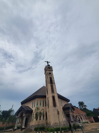 A low-angle shot shows a building and a cloudy sky. The building is a beige and brown, likely a church based on the tower topped with a cross. The steeple is light brown and extends into the sky. The building has a gabled roof and tall windows. The sky is overcast, featuring light and dark gray clouds. The location is Neni, Nigeria. Some nearby buildings are visible to the right of the frame. A palm tree grows near the church.
