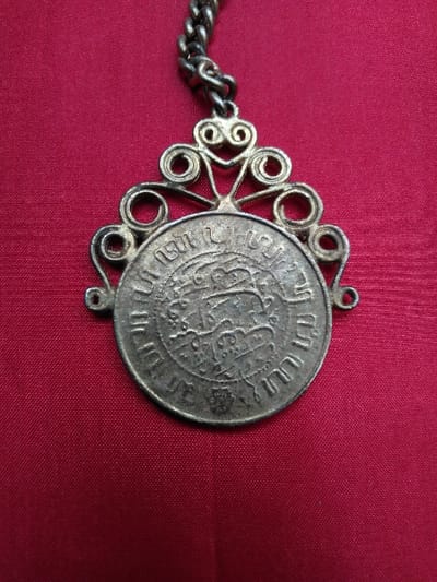 A tarnished metal pendant, located in Kaplice, Czechia, is the primary subject, displayed against a dark red, textured fabric background. The pendant comprises a circular medallion integrated into an ornate, openwork frame. The medallion's surface features raised, intricate script in an unidentified language or dialect, arranged in a circular pattern around a central, complex emblem. Both the script and emblem exhibit signs of wear, patination, and general surface degradation. The frame, composed of the same dull, grayish metal, is characterized by symmetrical scrollwork extending upwards from the medallion's top edge, forming a decorative crest. A short segment of a linked metal chain, consisting of interlocking oval links, is attached to a central loop at the apex of the frame. Two additional, smaller circular attachment loops are present on the lower sides of the frame, adjacent to the medallion. The entire metal piece shows discoloration, pitting, and tarnishing consistent with age. No individuals, actions, or other objects are visible in the scene. The illumination is diffused and even.