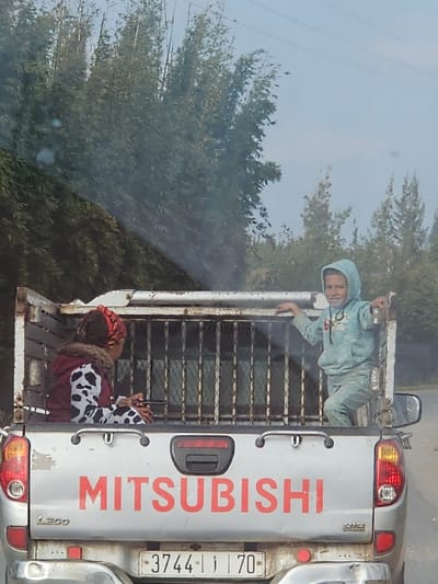 The image depicts the rear view of a silver Mitsubishi L200 pickup truck on a road in the city of Ameur عامر, Morocco. The truck's tailgate prominently displays "MITSUBISHI" in red lettering and a Moroccan license plate reading "3744 | 1 | 70". The truck bed is equipped with a metal cage structure.
Inside the caged bed, two individuals are present. On the left, an adult individual is seated on the bed floor, facing away from the viewer. This individual wears a patterned head covering, a dark jacket with a fur collar, and a white and black cow-print long-sleeved top. On the right, a young child stands, leaning against the metal cage and smiling towards the camera. The child is dressed in a light blue hooded tracksuit and holds a small, dark object in their right hand.
The background consists of dense green foliage, specifically tall, slender trees, under a clear light blue sky, indicating an outdoor, vegetated environment.