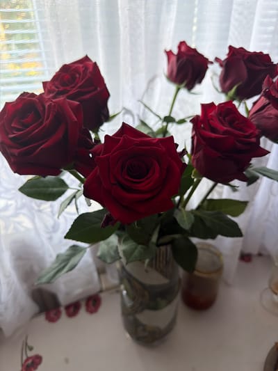 A close-up, top-down view captures a bouquet of deep red roses. The roses are the central focus, with their velvety petals, some fully bloomed and others still tightly furled. Green leaves and stems are visible amongst the flowers, suggesting a natural arrangement.
The background consists of sheer white curtains, diffusing soft light and creating a gentle, slightly blurred backdrop. Through the curtains, hints of an outdoor scene can be perceived, possibly indicating a window. On the right side of the image, a white surface, possibly a table, is visible. It is adorned with a few small, red floral motifs, and a glass jar containing an amber-colored liquid, likely honey or jam, is placed on it. A glimpse of a decorative plate with a floral pattern is also present.
The lighting suggests a bright day, likely daytime, with the light filtering through the window. The overall mood is one of natural beauty and domestic tranquility. No people are visible in the image. There is no visible text. The scene is likely set indoors, near a window, in a home environment. The overall impression is one of simple elegance.
