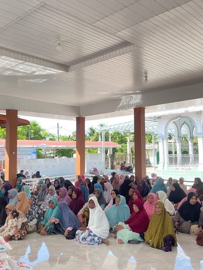 A large group of women, mostly elderly and wearing hijabs or head coverings of various colors, are seated in neat rows on a shiny, tiled floor in what appears to be a covered outdoor or semi-open area. Several young children are also present among the adults. The women are barefoot and generally face towards the right side of the frame, suggesting they are attending an event or listening to a speaker. Their expressions range from attentive to smiling, with some looking directly at the camera.

The setting is bright and sunny, indicated by clear shadows and strong daylight. The covered area has a distinctive white slatted ceiling with horizontal and diagonal patterns and visible light fixtures. Tall brown pillars support the roof structure. Beyond the seated women, in the background, elements of an ornate white building with arched gateways and railings, possibly a mosque, are visible, along with lush green trees and parts of other structures. Motorcycles are parked further back.

On the floor in the foreground, there are several white and blue sacks or bags with text, hinting at some form of distribution or community event. Visible text from banners and signs in the background includes "BID KEMUKIMAN BEURARO," confirming the location context of Aceh, Indonesia, where "Kemukiman" is an administrative unit. Other discernible words include "DENGAN RINGAN & NYAMAN," "UNTUK SERQURBA," "AT DICICIL," "PAYA," "Pulen," and "Tangise Pulen," possibly related to services, products like rice ("Pulen"), or charitable activities. The reflective floor shows clear reflections of the people and the ceiling structure.