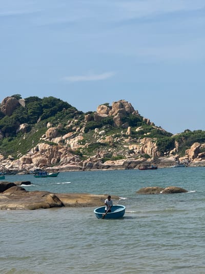 In the city of Thuận Thành, Vietnam, a calm blue sea stretches towards a rocky, green-covered hillside under a clear sky. A man in a light-colored shirt rows a small, round blue boat with a paddle in the foreground. Several other boats are visible further out in the water, including a teal-colored boat and a red and black boat closer to the shore. The rocky hill in the background is dotted with green vegetation.
