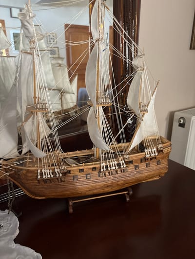 A detailed wooden model of a historical sailing ship is displayed on a dark wooden surface. The ship has multiple masts with white sails rigged with numerous thin ropes. The hull is intricately constructed with visible planking and rows of small square windows. The ship is supported by a wooden stand. In the background, a large mirror reflects the ship and the surrounding room. The environment appears to be an indoor setting, possibly a home or museum, with muted lighting. There are no people visible in the image. The location in the city of Ilion, Greece, is not discernible from the visual information provided.