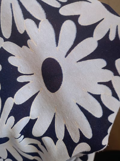 A close-up image depicts a piece of fabric featuring a bold, graphic floral pattern. The primary subject is this textile, which is adorned with stylized white daisy-like flowers against a very dark blue or black background. Each white flower shape has a distinct, dark circular center.
The fabric itself appears to be a woven material, possibly cotton or canvas, with a noticeable texture visible in the detailed crops. The white areas of the pattern are a light, off-white or cream color, and in some sections, a very faint, thin yellowish or light beige line outlines parts of the white petals, possibly from the weave or slight discoloration. The fabric is not perfectly flat, displaying gentle folds and creases, indicating it is likely draped or rumpled.
No specific activity or event is taking place; the image is a static portrayal of the patterned cloth. There are no people or other distinct objects visible beyond the fabric. The background is out of focus and indistinct. There are no discernible location cues within the image itself, though the provided context places it in Koghb, Armenia. No time of day, weather conditions, emotions, or visible text can be identified from the photograph.
