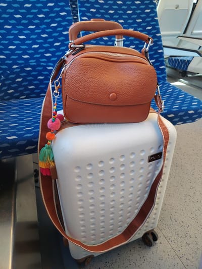 A brown leather handbag with a colorful tassel charm sits atop a white, textured suitcase on a seat in a public transportation vehicle, likely a train, given the patterned blue upholstery of the seats visible in the background. The handbag has a zippered compartment and a flap with a button closure. The suitcase has a prominent "KONO" logo badge and is equipped with wheels. The environment appears to be an interior seating area with a metallic railing to the left and a tiled floor below. The lighting suggests an indoor setting.