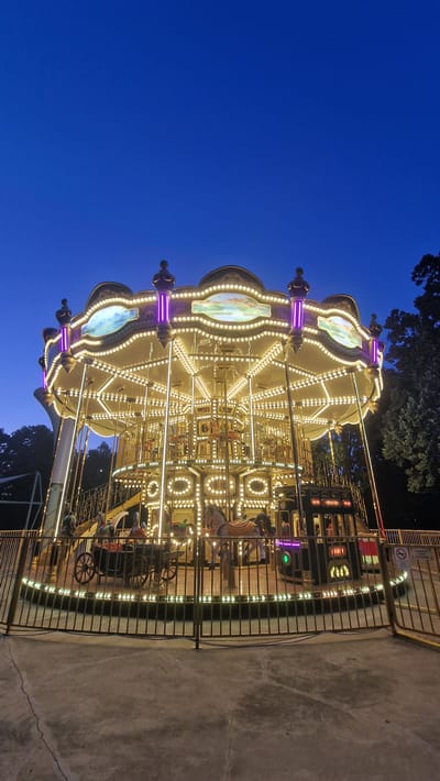 This image shows a carousel at dusk in an outdoor park setting. The carousel is illuminated with numerous warm white lights and some purple accent lights, highlighting its ornate design, which includes decorative elements and painted panels. Several carousel animals, primarily horses, are visible, some with riders. The carousel is surrounded by a metal fence, and a concrete pathway is in the foreground. Trees are visible in the background, and the sky is a deep blue, indicating the time of day is early evening or twilight. The scene appears to be peaceful and well-maintained, evoking a sense of amusement or nostalgia. The question of Keszthely, Hungary is not supported by the image content.