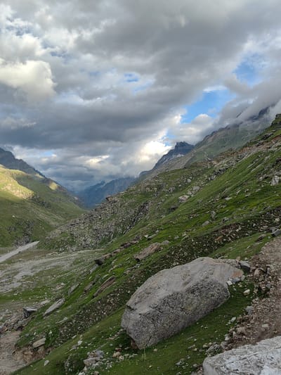 This is a vertical, eye-level, medium shot of a mountain valley in Yari-Khoksar, India, on a cloudy day. The valley floor is a gravelly riverbed, with a light-colored river running along the left side. The slopes of the valley are covered in green grass and scattered rocks, with a few larger boulders visible. The mountains in the distance are rugged and snow-capped, partially obscured by clouds. The sky is filled with a mix of dark gray and white clouds, with patches of blue sky visible. The overall impression is one of natural beauty and ruggedness. There are no people or animals visible in the image.