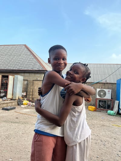 Two children, a boy and a girl, are embracing tightly. The boy is wearing a white tank top with blue trim and rust-colored corduroy pants. The girl is wearing a white top. An air conditioning unit is visible on a building in the background. The setting appears to be an outdoor area, likely in Iddah, Nigeria. The ground is dirt and gravel. Various objects, including a yellow jerrycan and metal containers, are scattered around. The sky is visible above.