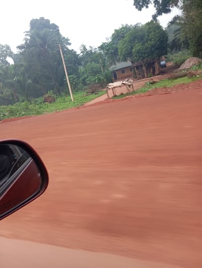 The image shows a blurred, low-angle view from a vehicle moving along a red dirt road. In the foreground, a portion of a red side-view mirror is visible on the left. The middle ground reveals a rural landscape with lush green vegetation and trees. A modest, light-colored building with a dark roof and windows is situated amidst the greenery, with a small blue vehicle parked nearby. A wooden structure, possibly a shelter or stall, stands in front of the building. In the background, more trees and foliage are visible, suggesting a tropical environment. A utility pole is also present, with wires extending from it. The overall impression is one of a journey through a remote, possibly agricultural, area in Nigeria.