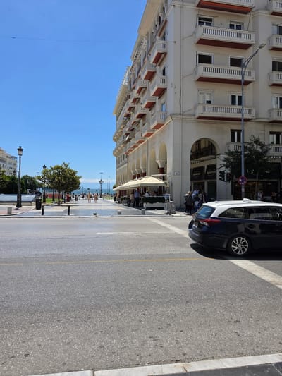 The image depicts a sunny day in Thessaloniki, Greece, likely on a waterfront promenade or a major street adjacent to it. In the foreground, a dark blue and white car, possibly a taxi, is parked on the right side of the road, facing away from the camera. The car has a distinctive white light on its roof. Across the road, a wide pedestrian area is visible, with people walking and sitting at outdoor cafes under awnings.

Dominating the right side of the image is a large, cream-colored building with multiple stories, featuring numerous balconies with red awnings and white railings. Arched walkways are visible at the ground level, housing shops and cafes. The street is paved with asphalt and marked with yellow and white lines.

To the left of the pedestrian area, there is a green space with trees and more people. A lamppost stands near the curb in the left foreground. In the background, the bright blue sky is visible, with some distant buildings and what appears to be the sea or a large body of water. The overall scene suggests a vibrant urban environment in a Mediterranean city, with a mix of architectural styles and daily life activities.