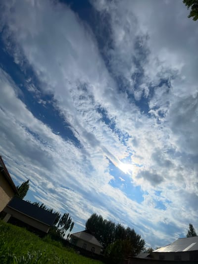 This is a low-angle, tilted shot of a cloudy sky over a rural landscape in Кызыл-Тоо, Kyrgyzstan. The sun is partially visible through thin, wispy clouds, creating a bright halo effect and a rainbow arc. Streaks of white and gray clouds stretch across a bright blue sky.

In the lower right quadrant, the scene transitions to ground level. A green grassy area is visible in the foreground. Behind the grass, there are several buildings with dark roofs and light-colored walls, characteristic of rural dwellings. Trees with dark green foliage are scattered throughout the scene, some appearing tall and slender, others more bushy. The overall impression is of an outdoor scene on a partially sunny day. The image appears to have been taken in the daytime, likely late morning or early afternoon, given the brightness of the sun. There is no visible text in the image.