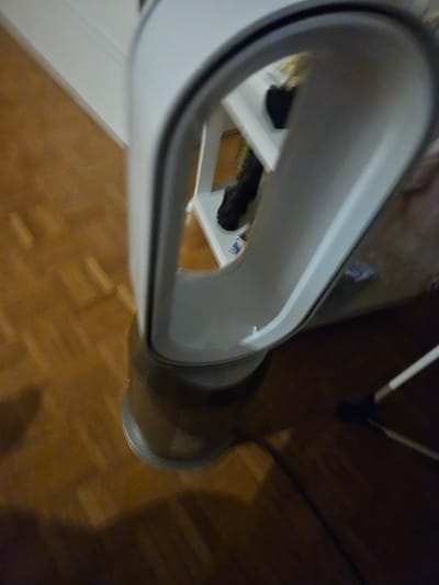 A white and grey bladeless fan is positioned on a wooden floor, partially obscuring a white shelf. The fan’s looped structure is prominent, and its base, a grey cylindrical unit with a perforated texture, is visible. A black cable extends from the fan's base and trails across the floor towards the lower right corner of the image. The floor displays a parquet pattern with variations in wood grain and color. In the background, a sliver of a white wall is visible. The scene is indoors, and no specific location in Versailles, France, can be identified from the visual information provided.