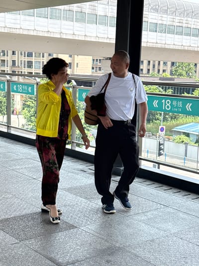 The image captures two older individuals, a man and a woman, standing on a light grey tiled surface, likely an elevated walkway or a station platform in an urban setting. The scene is brightly lit, indicating daytime with clear weather.
The man, on the right side of the frame, is wearing a white short-sleeved t-shirt, dark trousers, and dark blue sneakers. He has short hair and is looking downwards. A dark brown shoulder bag hangs from his shoulder.
The woman, positioned to the left of the man, is dressed in a bright yellow long-sleeved jacket over a floral-patterned ensemble of top and pants, featuring maroon and darker hues. She wears black and white flat shoes, and a light-colored straw hat is visible on her head. A light green bracelet adorns her left wrist. She appears to be looking upwards or to the side.
In the background, modern buildings with multiple windows are visible. Beyond the immediate walkway, green foliage and glimpses of a street scene, including a traffic light and road signs, can be observed. Two prominent green signs with white text display "18 号线" (Line 18) and "Line 18" in English, strongly indicating the proximity to a metro or subway station. Other visible text includes "40" on a small sign, likely a speed limit, and a red circular sign with a blue background and a red diagonal line, signifying "no stopping" or "no parking."
The setting is an urban environment, consistent with public transport areas in cities like Tiaodeng, China, as suggested by the visible Chinese text. The two individuals appear to be together, either walking or waiting.