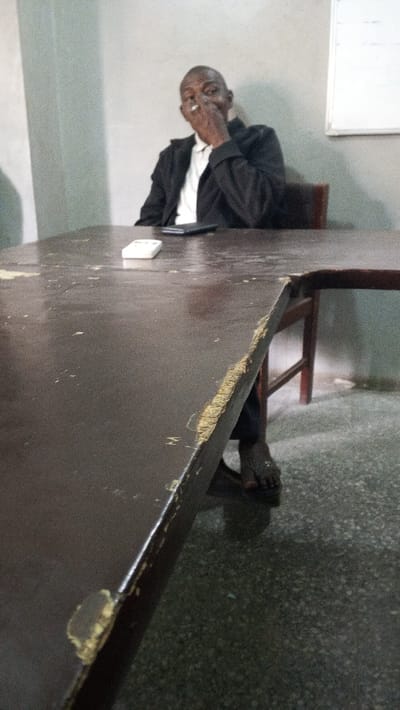 The scene is an indoor environment in Dakata, Nigeria. An adult male with dark skin is seated at a dark brown, heavily worn table. He is dressed in a white collared shirt and a dark blazer. His right hand is raised to his face, holding a white cylindrical object, consistent with a cigarette, between his fingers. A white rectangular object, resembling a remote control, and a dark smartphone are positioned on the table surface in the foreground. The table shows significant wear, with multiple chips, scratches, and areas where the laminate is peeling, exposing lighter material beneath. The man's left foot, bare and wearing a dark sandal, is visible under the table. The background features plain, light-colored walls and a dark, speckled floor. A white rectangular board is visible on the wall to the right of the man.