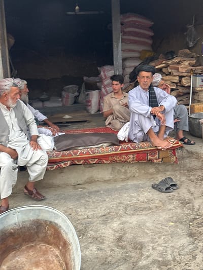 In Chaman City Tehsil, Pakistan, a group of men are gathered outdoors. The scene appears to be a shop or marketplace, with stacks of goods, including sacks of what looks like flour or grain, and piles of wood in the background. In the foreground, four men are visible. Two older men are seated on the left, wearing traditional Pakistani attire, including vests and loose-fitting trousers, and patterned caps. One of them has a white beard. In the center, a younger man in a light brown tunic is seated, looking to his left. To his right, an older man in a light blue tunic and a black scarf around his neck is seated cross-legged on a patterned rug. He is looking forward, and his hands are clasped in his lap. A pile of sacks is visible behind him, along with some wooden planks. To the right of the rug, a pair of black sandals is on the ground, and further to the right, another person's leg and foot are visible, wearing black sandals. A large, weathered metal basin is in the bottom left of the image. The ground is rough and concrete-like.