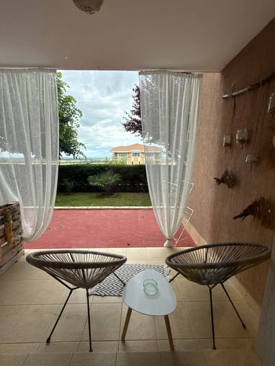 The image depicts a covered outdoor patio area with a view of a garden and distant buildings. In the foreground, two distinctive modern chairs with woven seating and thin metal legs are positioned on a tiled floor. A small, oval white coffee table with wooden legs sits between the chairs, and an ashtray is visible on its surface. A patterned rug is partially visible beneath the coffee table and the chair on the left. White sheer curtains hang from the ceiling on either side of the view, framing the outdoor scene.

The outdoor area features a red-tiled floor and a garden with a neatly trimmed hedge. Beyond the hedge, a small palm tree is planted. In the background, a two-story building with an orange roof is visible, along with a landscape of fields and a cloudy sky. The wall of the patio on the right side is textured and painted a light brown, adorned with several decorative items, including what appear to be jars hanging by twine and two rustic fish-like figures made of wood. A metal drying rack is visible behind the curtain on the right.

The scene suggests a tranquil setting, possibly a residential patio or a small guesthouse, located in Kosharitsa, Bulgaria, offering a view of the surrounding countryside. There are no people visible in the image.