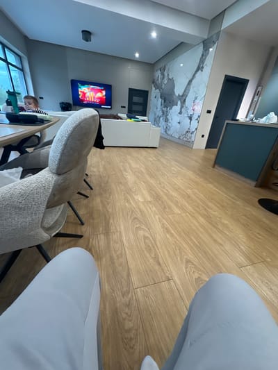 The image captures a modern interior space, likely a living room or open-plan area within a residence in Zhelyazno, Bulgaria, presented from a tilted, low-angle perspective. The predominant flooring consists of light-colored wooden planks.
A notable feature is a large accent wall in the mid-ground adorned with a striking white and grey marble-like pattern. Recessed spotlights are visible in the ceiling above this wall and in other parts of the room. To the right, a dark blue cabinet or counter contrasts with the lighter tones.
On a wall in the background, a flat-screen television displays colorful visuals, possibly from a video game or animated content. Faint text, including "BBC" and numbers like "10" and "20", can be discerned on the screen. Below the television, a window provides natural light, showing an overcast outdoor scene.
In the lower-left foreground, a young boy with short brown hair is seated at a table, engaged with a green object in front of him. Only his head and upper torso are clearly visible.
Furniture includes several chairs; one in the immediate foreground features light grey/beige textured fabric, while others have a lighter grey/blue fabric. A white sofa is positioned against a wall further back. Dark-colored doors are visible in various parts of the room. The overall atmosphere suggests a contemporary and functional living environment.