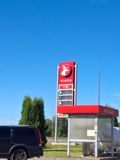 This media file captures a bright, sunny day in Glāžšķūnis, Latvia, with a clear blue sky dominating the scene. The primary focus is a red gas station sign displaying fuel prices. The sign prominently features the "VIADA" logo and the brand name "VIADA" in large white letters. Below, digital displays show fuel prices for different grades, including "MultiX" and what appears to be "95" octane. Other labels like "D" and "GAZE" are also visible, indicating different fuel types or services.
To the right of the main sign, a section labeled "AUTOGĀZE" suggests the availability of autogas. This area includes a glass enclosure with some equipment visible inside, possibly related to fuel storage or dispensing.
In the foreground, the rear of a dark-colored SUV is visible, parked at the gas station. The tire of the SUV has white lettering on its sidewall, with "INCEPTION" and "RIPAAX" being partially readable. Lush green trees form a backdrop behind the gas station, and a patch of grass is visible along the roadside. The overall atmosphere is one of a typical day at a fueling station, with no people or specific events depicted. The time of day appears to be mid-morning or afternoon due to the strong sunlight and clear sky.