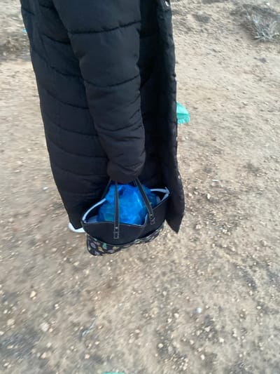 An overhead, first-person view captures a person walking outdoors on a sandy, dirt path. The ground is light brown, covered with small pebbles and sparse dry vegetation, suggesting an arid or semi-arid environment. The person's lower body is visible, dressed in a black padded jacket or sleeveless vest, which occupies the left side of the frame.
A black bag, possibly a tote or shoulder bag, is being carried on the right. It features black straps and a subtle, colorful pattern on its exterior. Inside the bag, a prominent, bright blue plastic bag is visible, suggesting contents. A small, light green fabric item or bag is partially seen near the black garment, close to the carried bag.
The blurred background implies movement, indicating the person is walking. The natural, bright lighting suggests it is daytime. The scene is consistent with an outdoor setting in a dry region, aligning with the provided location context of الغنادة, Tunisia. No text is visible in the image.
